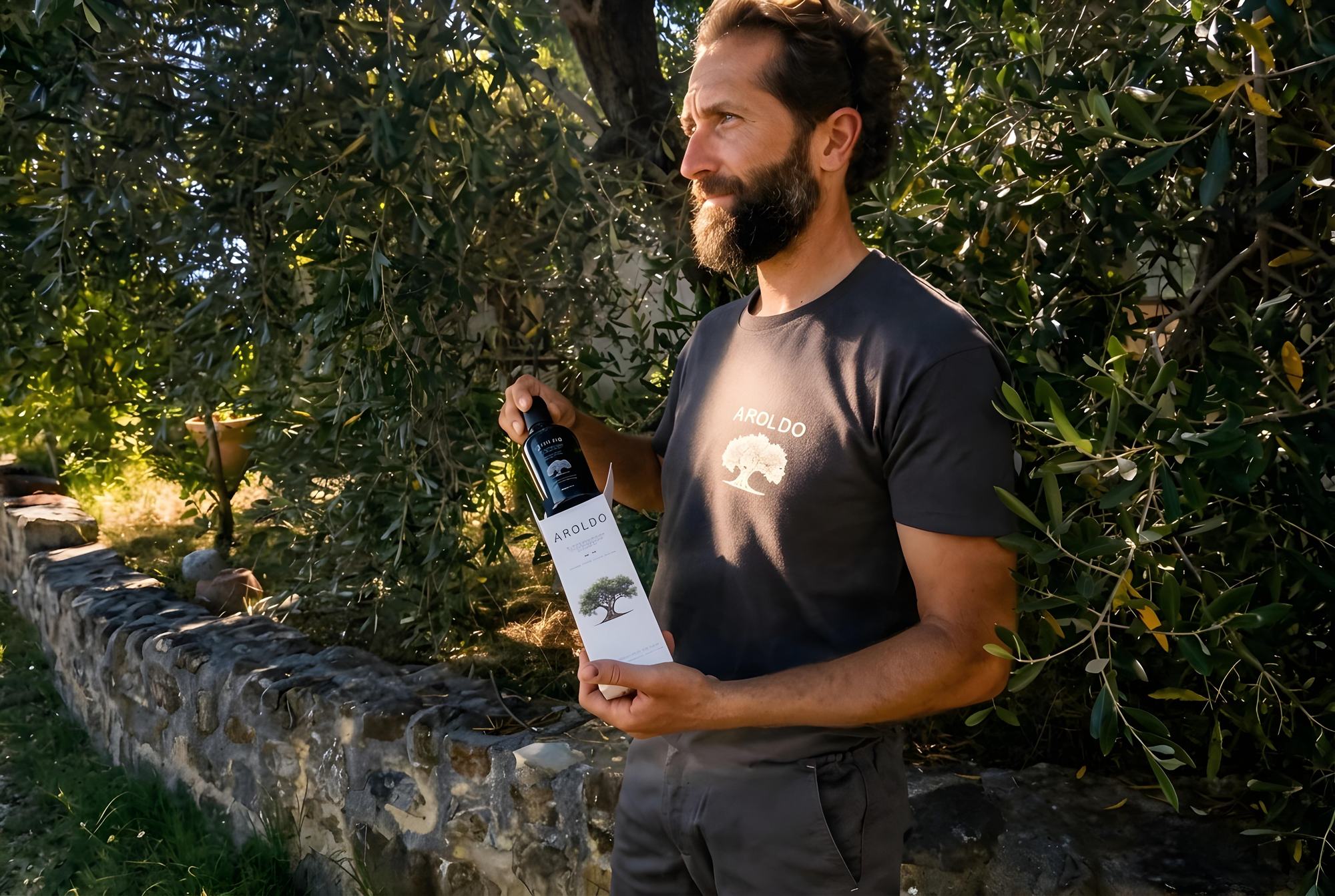 Gianluca tiene in mano una bottiglia d’olio tra gli ulivi, con la maglietta Aroldo, accanto a un muretto in pietra